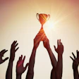 Silhouetted hands reaching up to grasp a golden trophy against a warm, glowing background