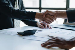 Two business professionals shake hands over documents lying on top of a desk.