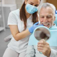 An older man smiles while looking into a mirror. He is looking at his new smile. A woman dentist sits next to him.