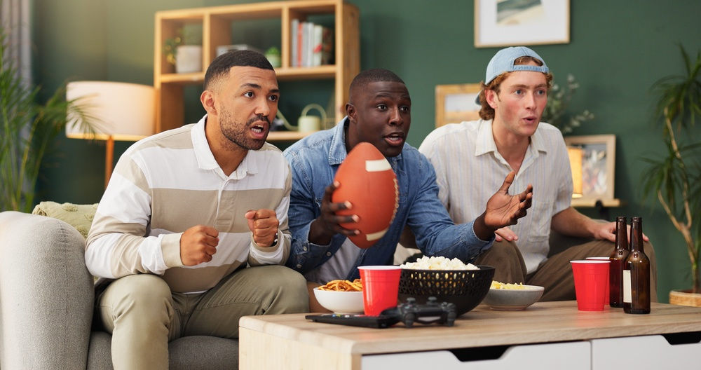 Three male football fans watch a game on a couch. They are surprised expressions on their faces. Nearby are bottles of beer, popcorn, and chips.