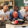 Three male football fans watch a game on a couch. They are surprised expressions on their faces. Nearby are bottles of beer, popcorn, and chips.