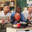 Three male football fans watch a game on a couch. They are surprised expressions on their faces. Nearby are bottles of beer, popcorn, and chips.