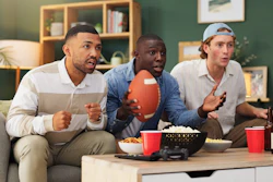 Three male football fans watch a game on a couch. They are surprised expressions on their faces. Nearby are bottles of beer, popcorn, and chips.