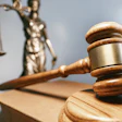 A wood gavel rests on a sound block with a brass Lady Justice statue in the background.