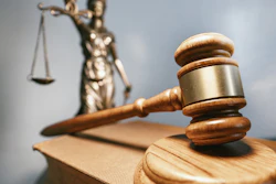 A wood gavel rests on a sound block with a brass Lady Justice statue in the background.