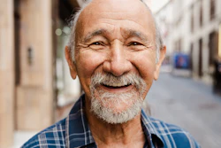 An older man with a beard smiles at the viewer. In the background, blurred, is a city street.