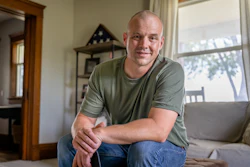 A man wearing an olive green T-shirt looks at the camera. Behind him is a folded American flag, indicating he is a veteran of the U.S. Armed Forces.