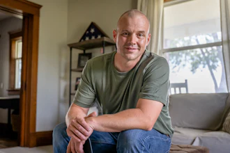 A man wearing an olive green T-shirt looks at the camera. Behind him is a folded American flag, indicating he is a veteran of the U.S. Armed Forces.