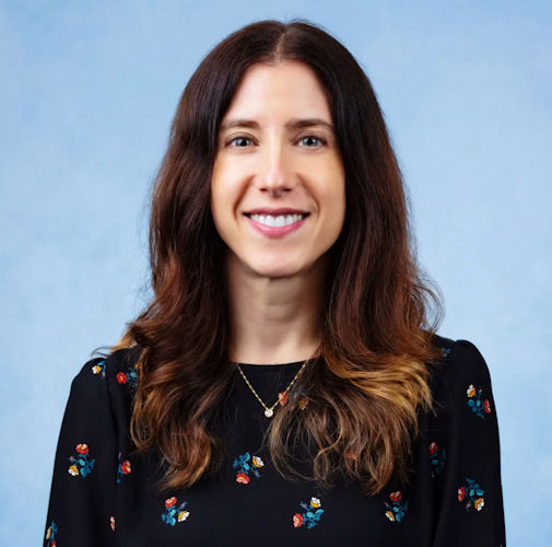Professional headshot of a woman with long dark hair wearing a black floral patterned top against a light blue background