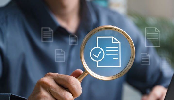 A man holds up a magnifying glass to a superimposed image of a document with a check mark pictured next to it. The image symbolizes a review and approval process.