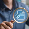 A man holds up a magnifying glass to a superimposed image of a document with a check mark pictured next to it. The image symbolizes a review and approval process.