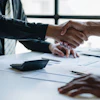 Two business professionals shake hands over a desk with documents and a calculator during a meeting.