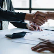 Two business professionals shake hands over a desk with documents and a calculator during a meeting.