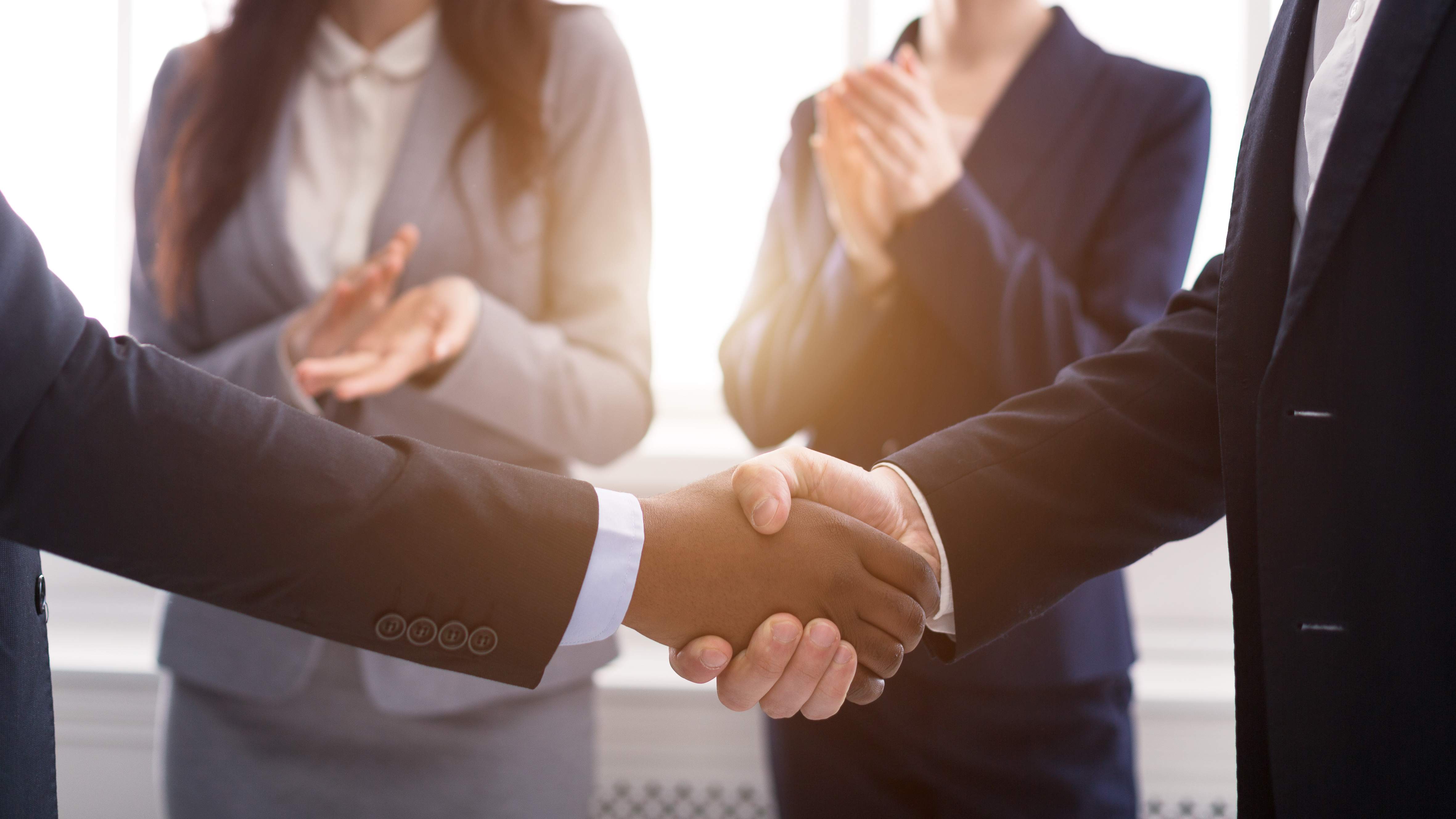 Business professionals shake hands with colleagues applauding in the background.