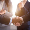 Business professionals shake hands with colleagues applauding in the background.