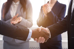 Business professionals shake hands with colleagues applauding in the background.