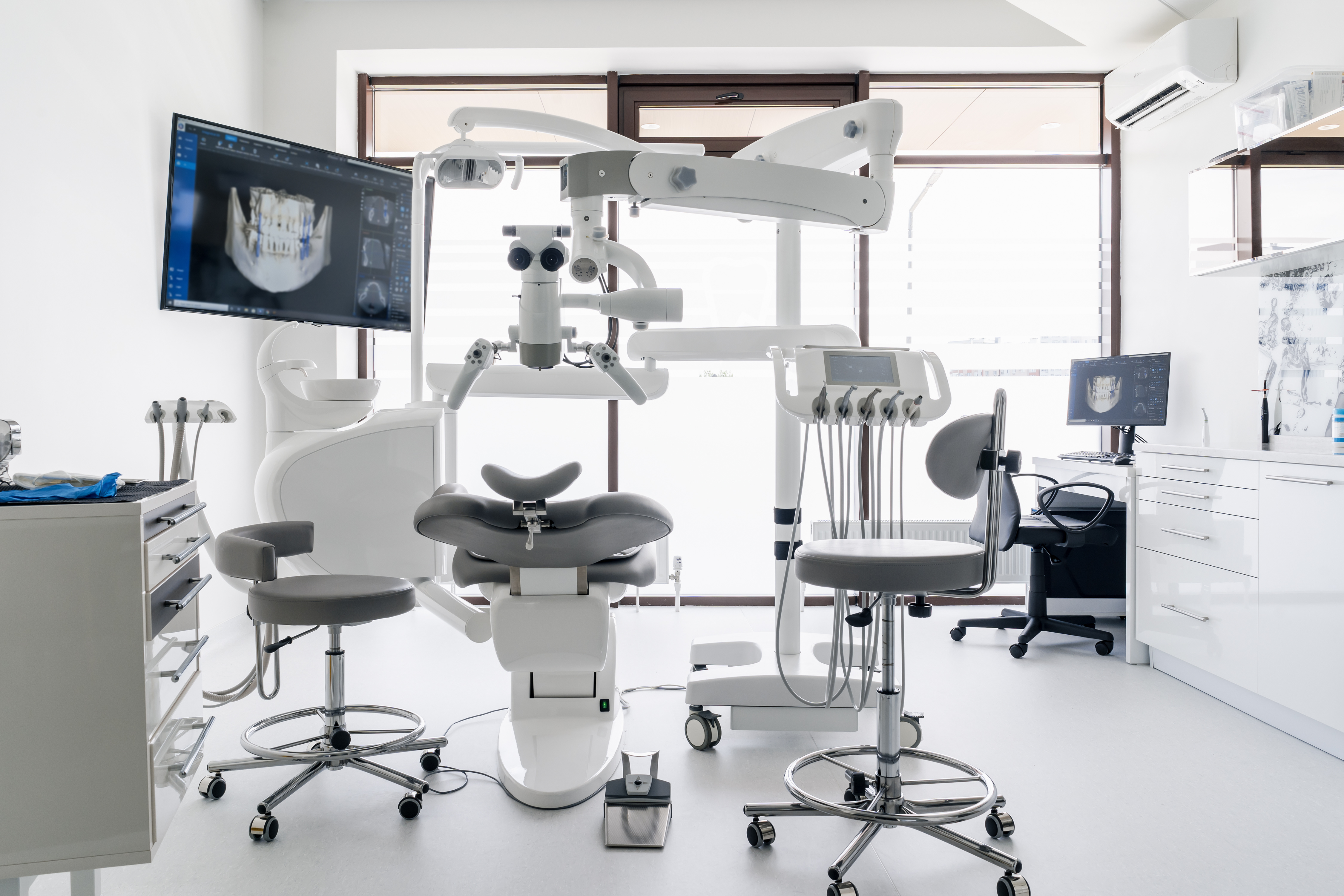 Modern dental operatory with treatment chair, overhead surgical microscope, digital imaging monitor, and professional equipment in a bright clinical setting.