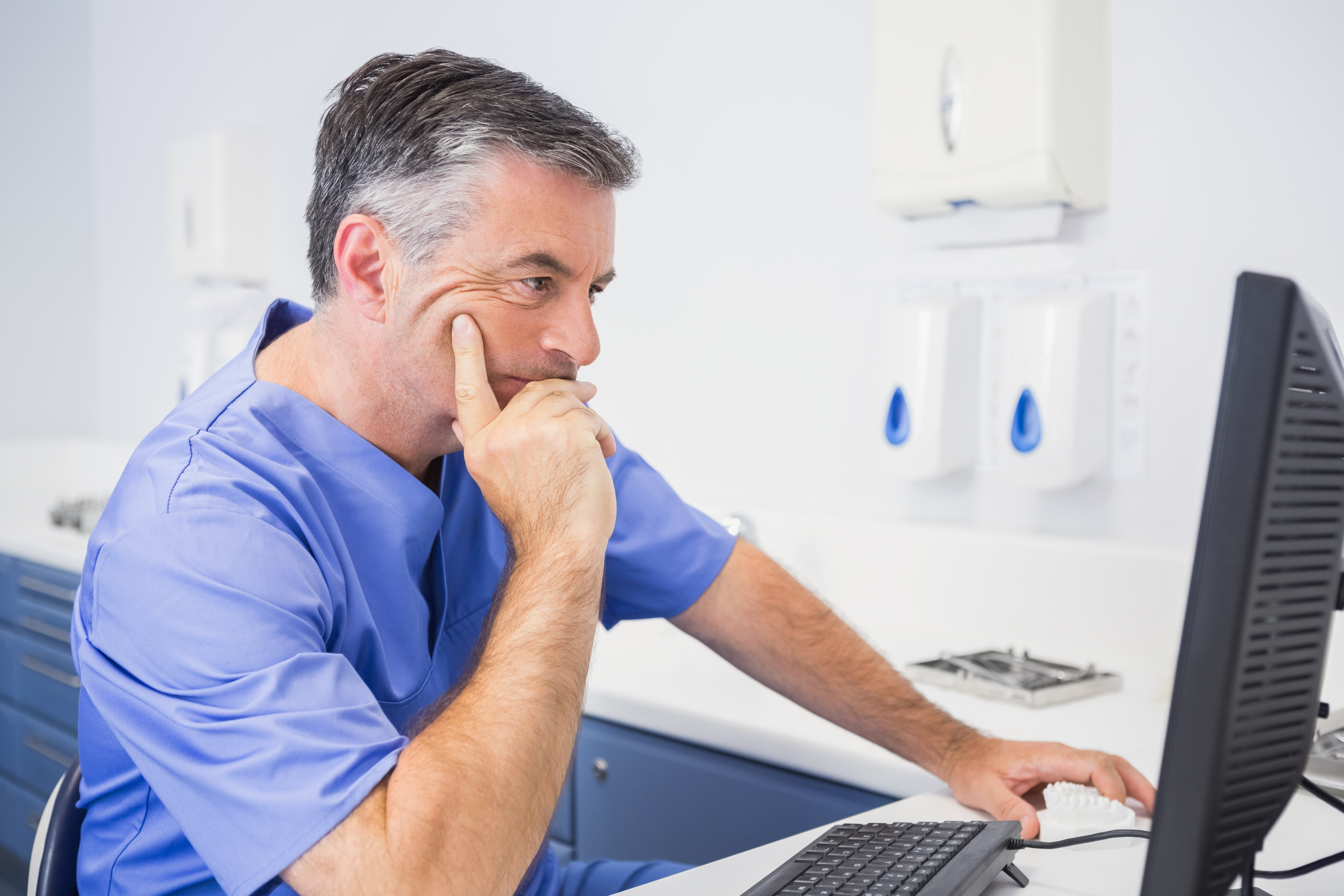 A male dentist wearing blue scrubs looks at a desktop computer.