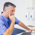 A male dentist wearing blue scrubs looks at a desktop computer.