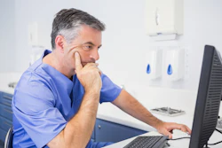 A male dentist wearing blue scrubs looks at a desktop computer.