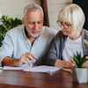 An older couple reviews paper on a kitchen table. Nearby are plants and a laptop.