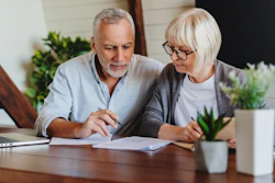 An older couple reviews paper on a kitchen table. Nearby are plants and a laptop.