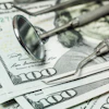 Dental tools, including a mirror and explorer, lie on top of several $100 bills, signifying cost of dental care.