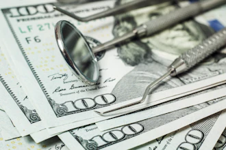Dental tools, including a mirror and explorer, lie on top of several $100 bills, signifying cost of dental care.