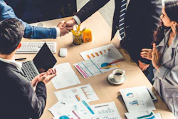Business team members shake hands over a table holding documents, computers, and charts.