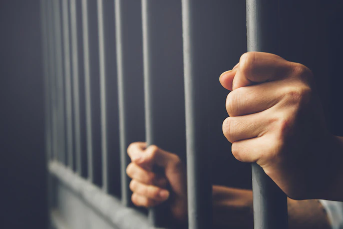Hands grip prison bars in dim lighting, symbolizing incarceration and confinement.