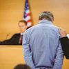 A man hangs his head down, with his hands behind his back, as he appears before a judge.