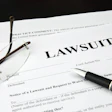 A white document is shown titled 'Lawsuit,' with glasses and a fountain pen resting on top.