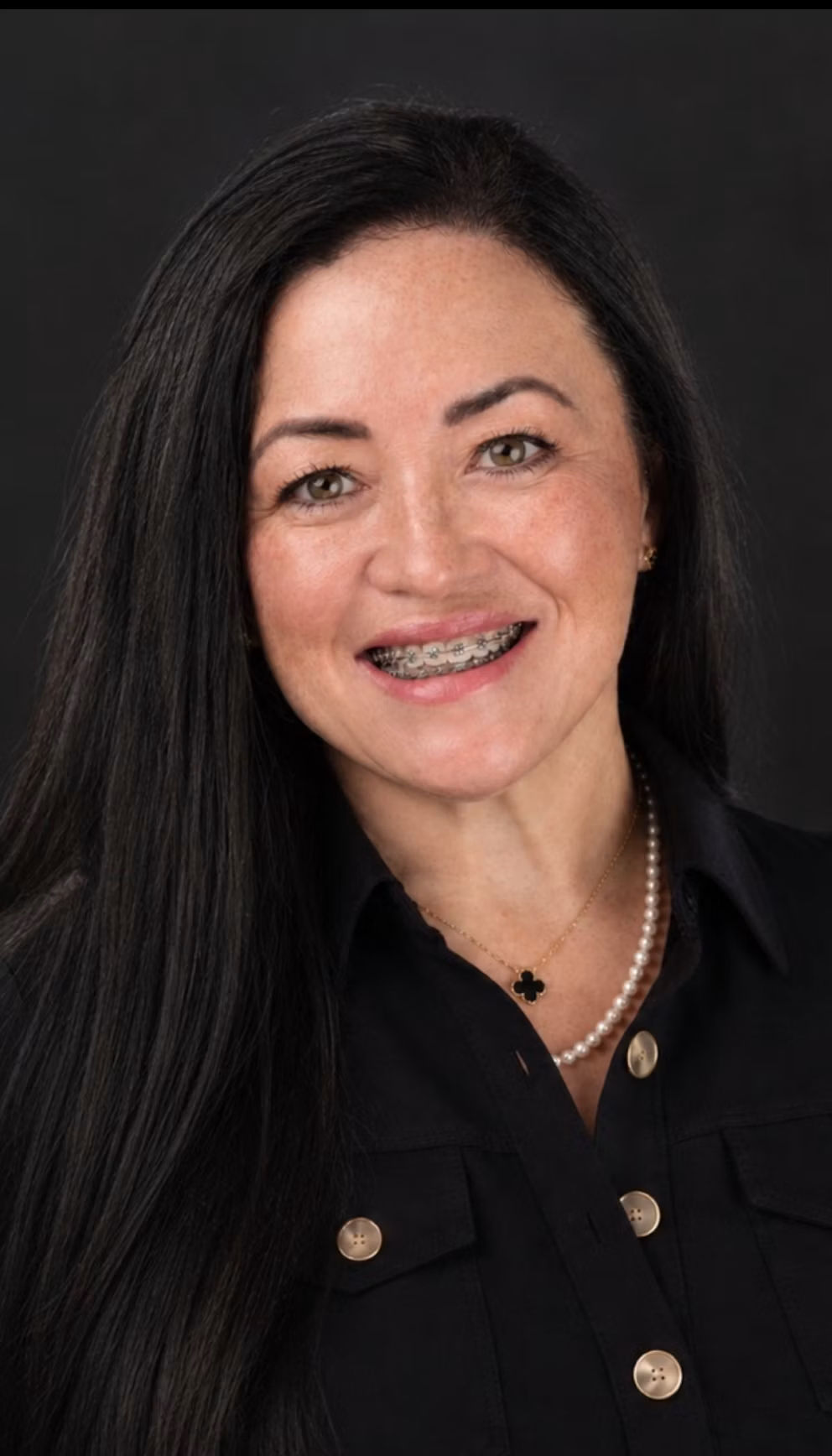Professional headshot of woman with long dark hair wearing black blazer and pearl necklace, smiling against dark background