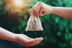 Two hands exchange a small fabric pouch in a sunlit garden setting, symbolizing giving.
