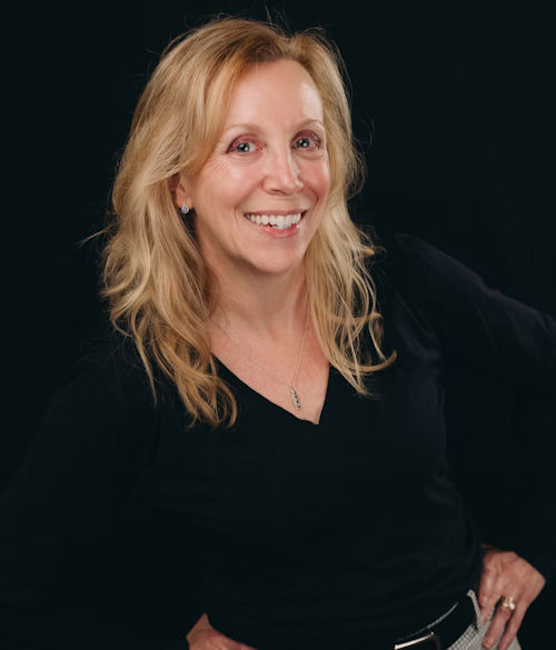 Professional headshot of a smiling woman with blonde wavy hair wearing a black top and pendant necklace
