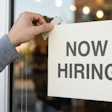 Close-up of a person taping a 'Now Hiring' sign to a glass door.