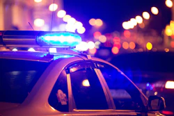 A police car is shown with its blue overhead lights illuminated at night with city lights in the background.