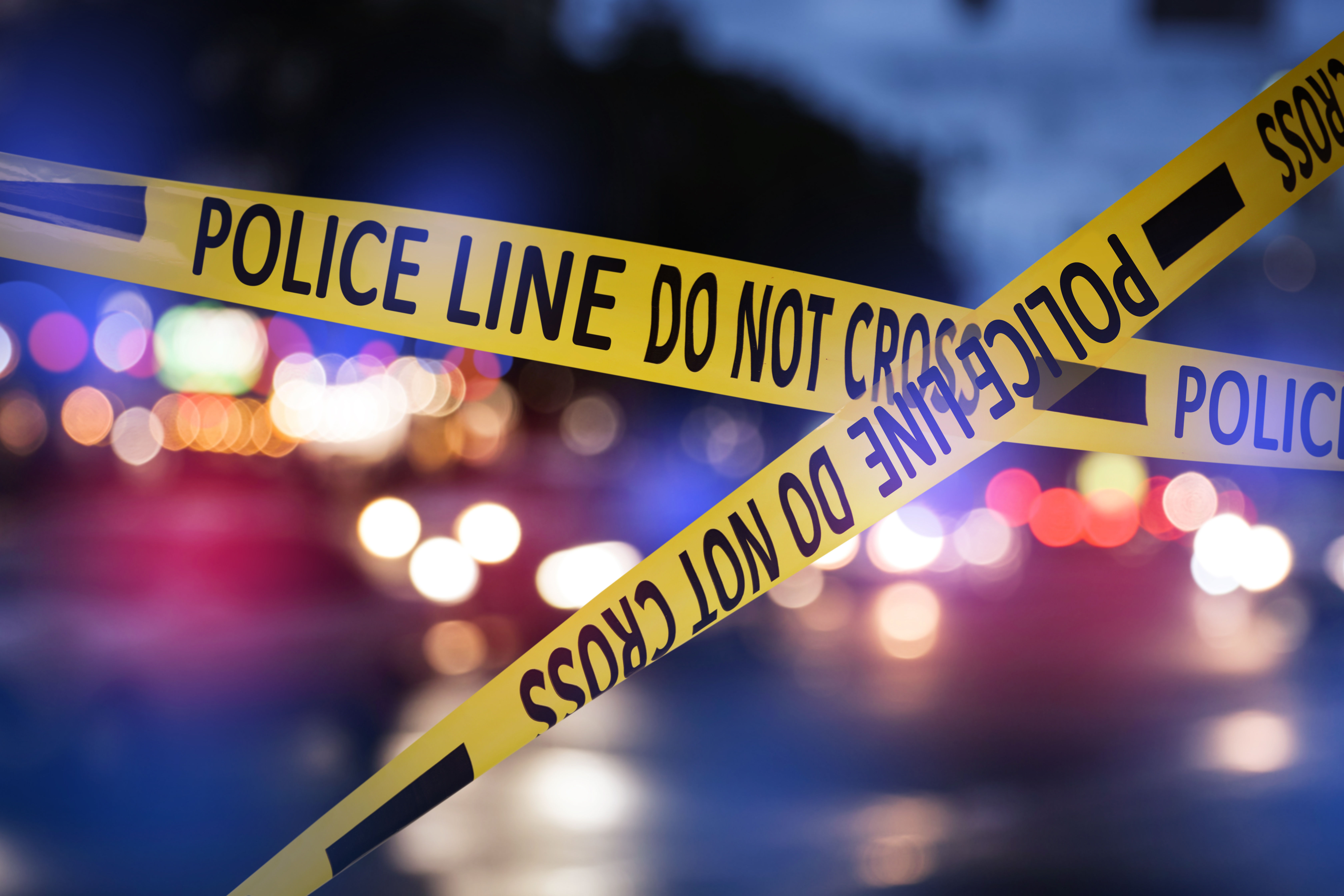 Yellow-and-black police crime scene tape crossing diagonally with blurred emergency vehicle lights in the background.