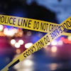 Yellow-and-black police crime scene tape crossing diagonally with blurred emergency vehicle lights in the background.