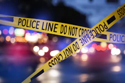 Yellow-and-black police crime scene tape crossing diagonally with blurred emergency vehicle lights in the background.