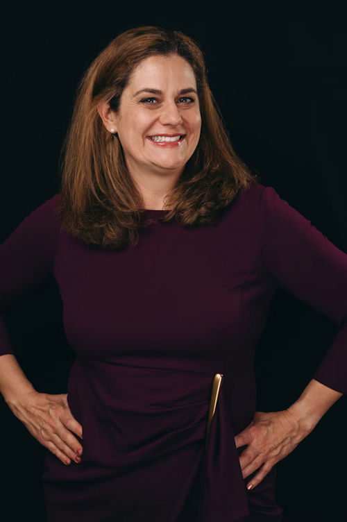 Professional headshot of a woman with shoulder-length auburn hair wearing a burgundy top against a dark background