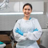 A woman wearing PPE smiles at the viewer. Behind her is a modern dental operatory.