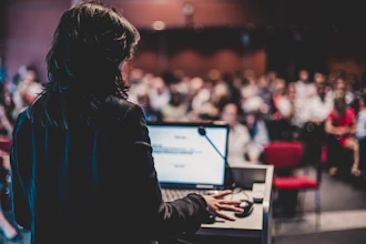 A woman speaker stands at a lectern addressing an audience, which is blurred.