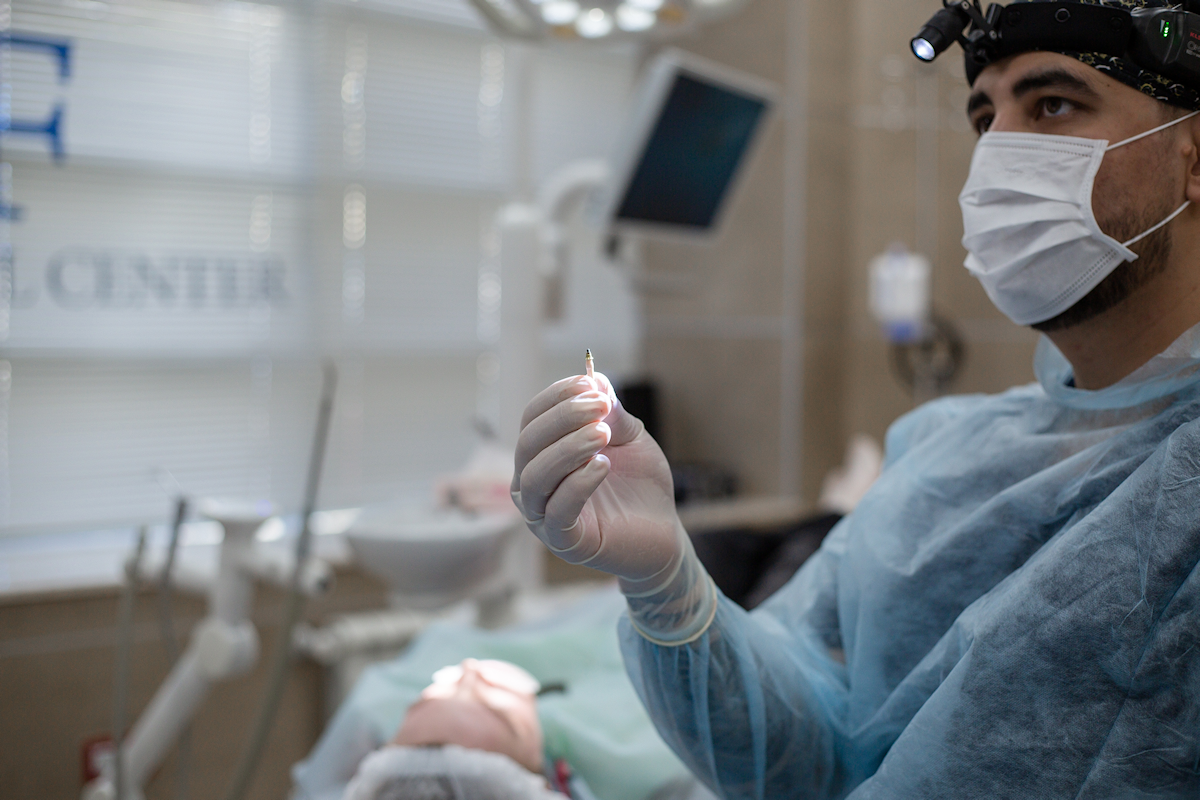 A dentist wearing a surgical mask and gloves holds a dental implant abutment.