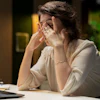 A woman working at a desk rubs her eyes. Behind her is a blurred office.