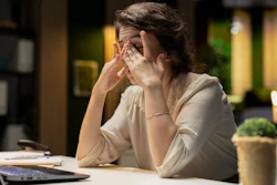 A woman working at a desk rubs her eyes. Behind her is a blurred office.