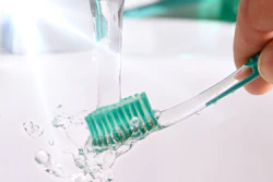A hand holds a green-bristle toothbrush under running water.