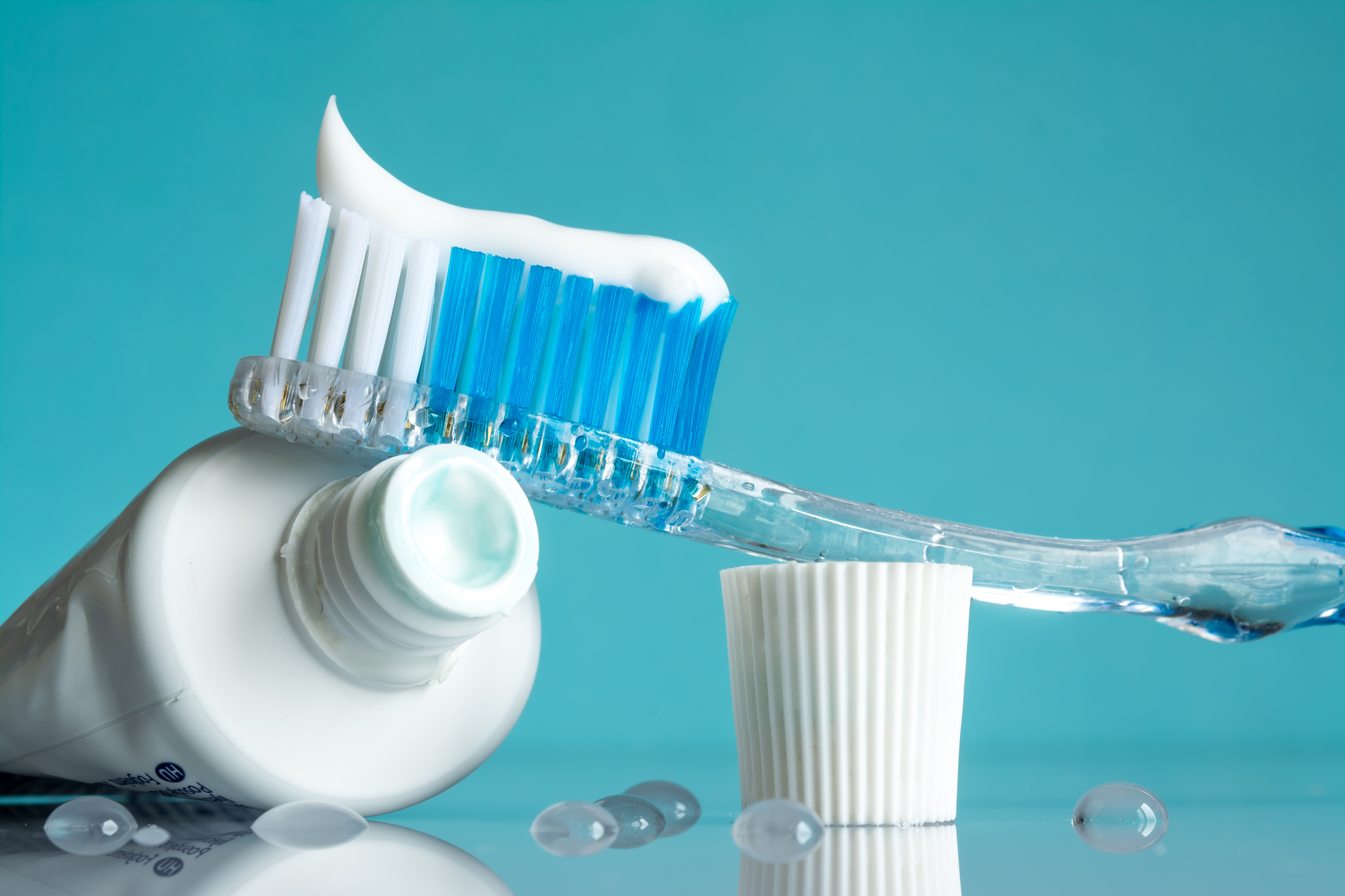 A toothbrush with white toothpaste and blue bristles is set next to a white cup on a turquoise background.