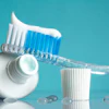 A toothbrush with white toothpaste and blue bristles is set next to a white cup on a turquoise background.