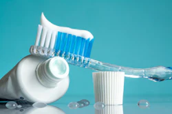 A toothbrush with white toothpaste and blue bristles is set next to a white cup on a turquoise background.